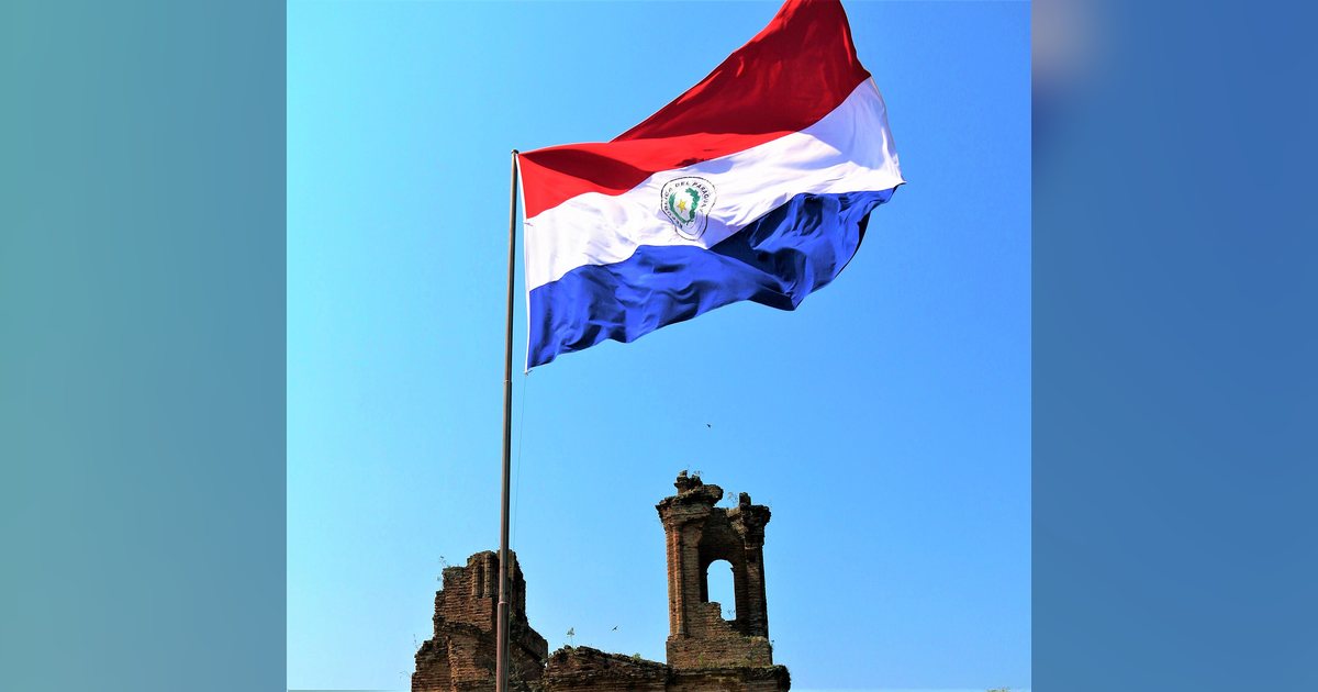 Bandera paraguaya con el escudo de la República en la franja blanca, ondeando junto a unas ruinas de ladrillo
