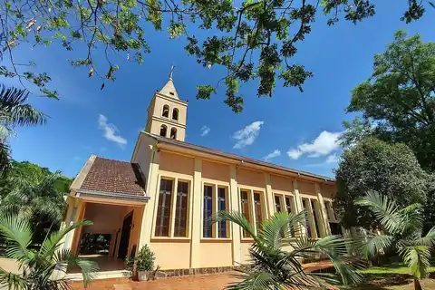 Iglesia de paredes color crema con campanario y ventanales altos, rodeada de palmeras y árboles bajo un cielo azul, en Juan León Mallorquín