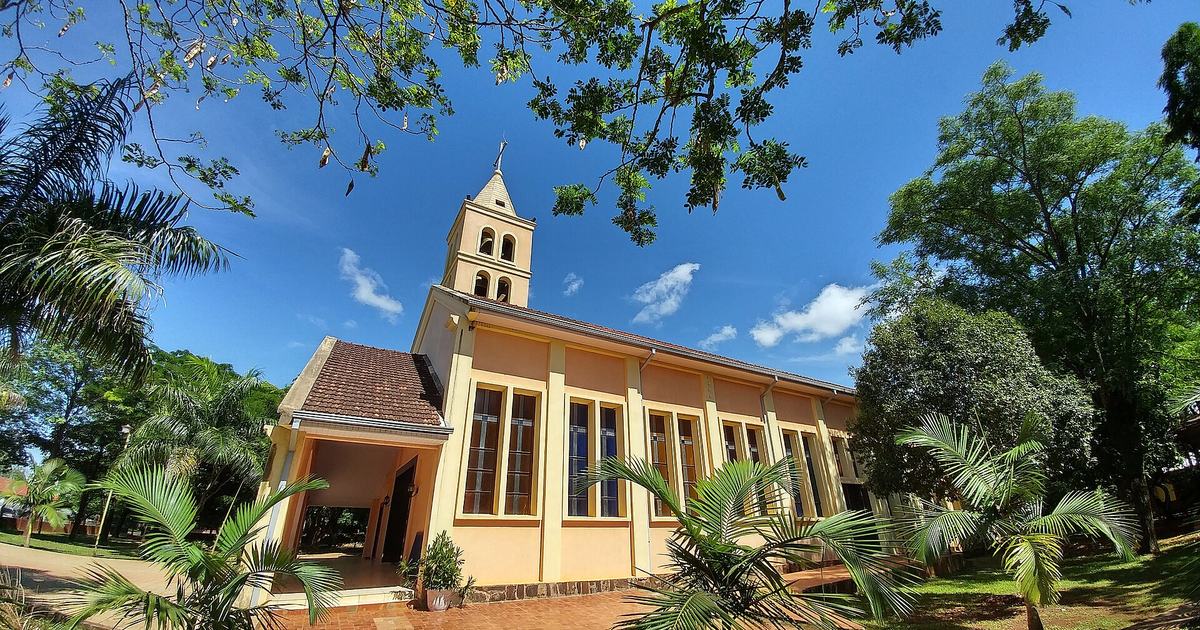 Iglesia de paredes color crema con campanario y ventanales altos, rodeada de palmeras y árboles bajo un cielo azul, en Juan León Mallorquín