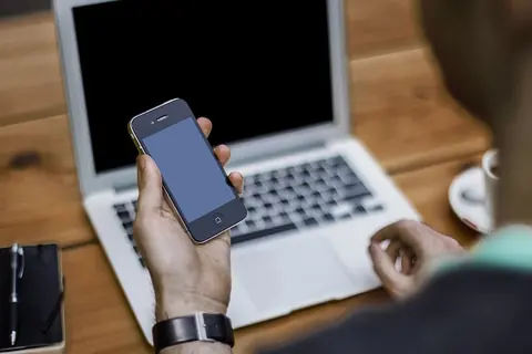 Foto: Alejandro Escamilla alejandroescamilla / Wikimedia Commons · CC0 Una persona sostiene un celular frente a una computadora portátil sobre una mesa de madera