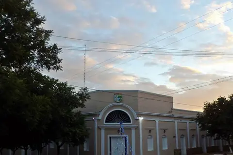 Edificio municipal de una planta con arco en la entrada y escudo en la fachada, entre árboles, al atardecer