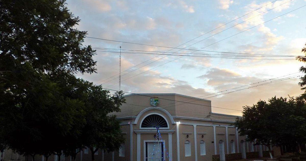 Edificio municipal de una planta con arco en la entrada y escudo en la fachada, entre árboles, al atardecer