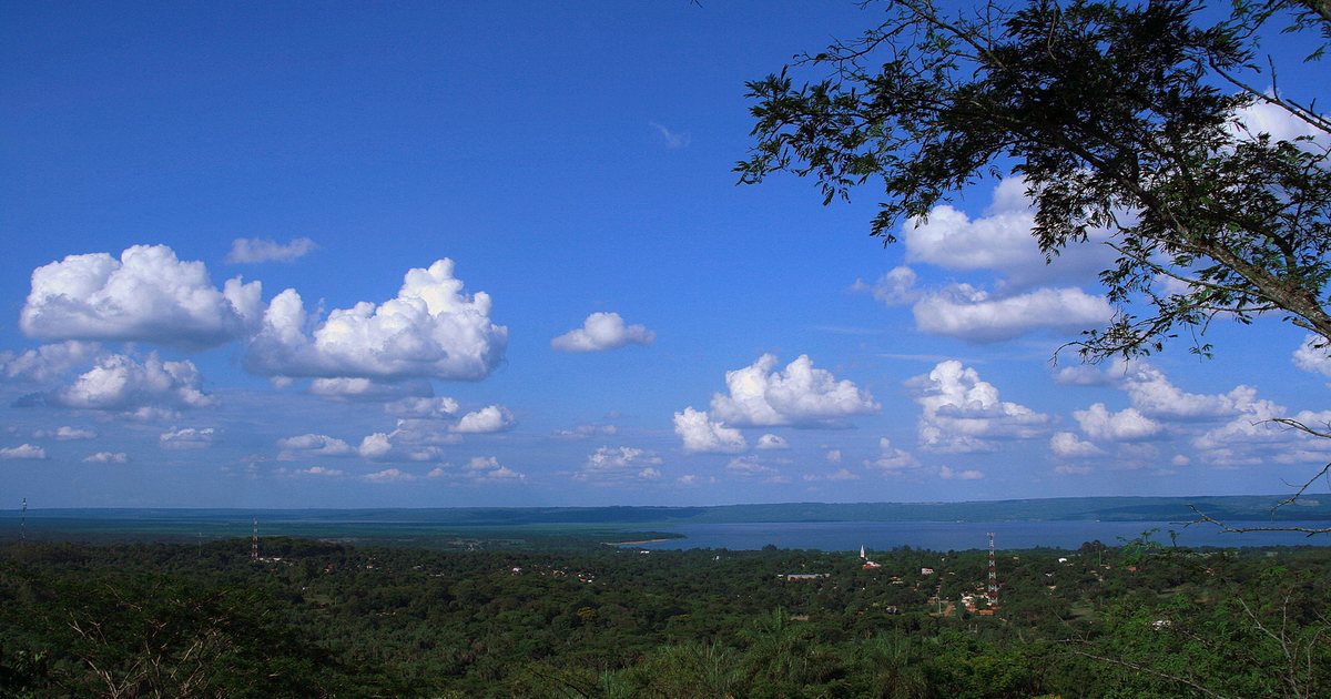 Vista desde lo alto de Areguá, con el bosque, algunas casas y el lago Ypacaraí al fondo, bajo un cielo azul con nubes