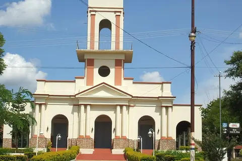 Iglesia blanca con detalles color salmón, campanario alto y tres puertas en arco, con un camino de piedra entre arbustos al frente