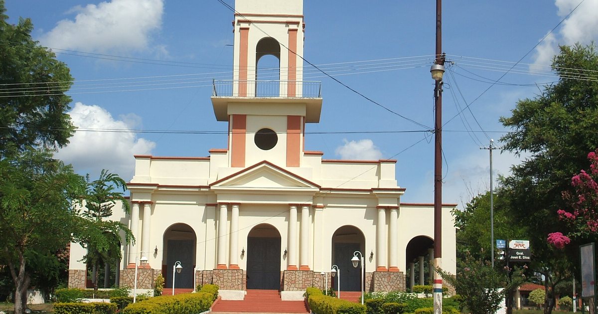 Iglesia blanca con detalles color salmón, campanario alto y tres puertas en arco, con un camino de piedra entre arbustos al frente