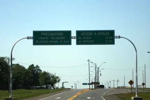 Pórtico con dos carteles verdes sobre una ruta asfaltada: uno avisa de un cruce peligroso y el otro indica el acceso a Ayolas, a 52 kilómetros