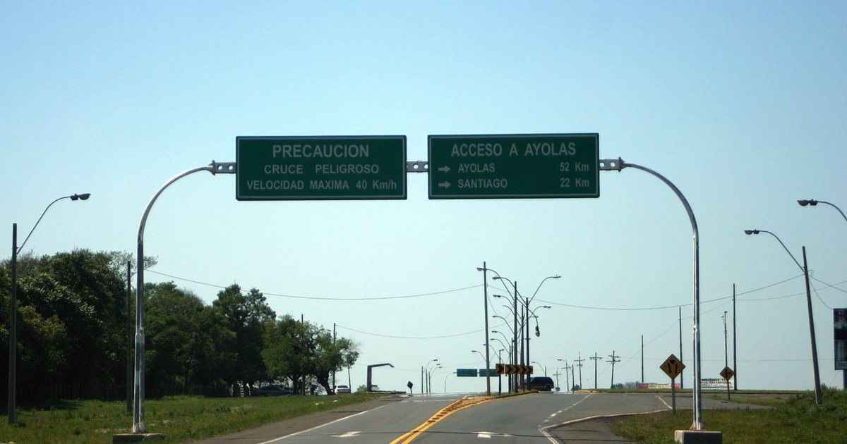 Pórtico con dos carteles verdes sobre una ruta asfaltada: uno avisa de un cruce peligroso y el otro indica el acceso a Ayolas, a 52 kilómetros