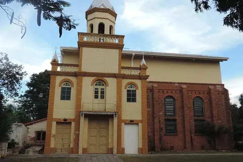 Foto: Rodney Damián / Wikimedia Commons · CC BY-SA 3.0 Fachada de una iglesia de ladrillo con frente amarillo y naranja, una torre con cúpula blanca y una cruz, al final de un camino de piedra entre árboles
