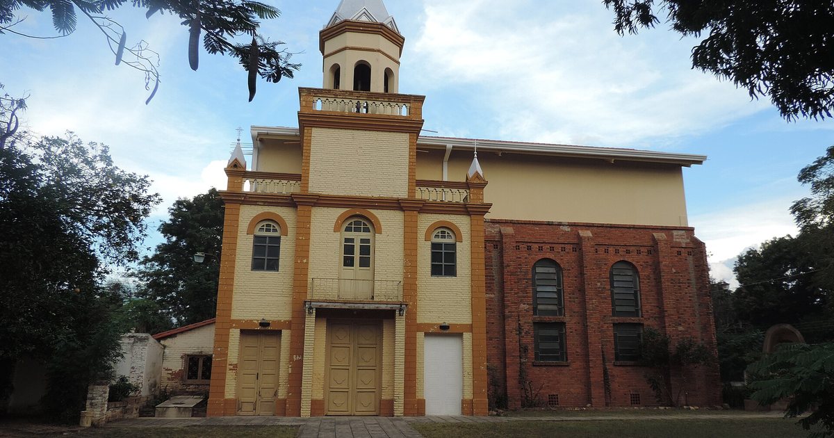 Fachada de una iglesia de ladrillo con frente amarillo y naranja, una torre con cúpula blanca y una cruz, al final de un camino de piedra entre árboles
