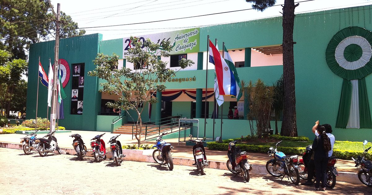 La sede de la Municipalidad de Caaguazú, un edificio verde con banderas al frente, motos estacionadas en la vereda y pinos a los costados
