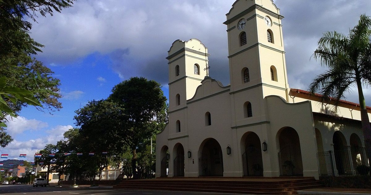 Fachada blanca de la catedral de la Inmaculada Concepción de Carapeguá, con dos torres y arcos, junto a la calle y bajo un cielo azul con nubes
