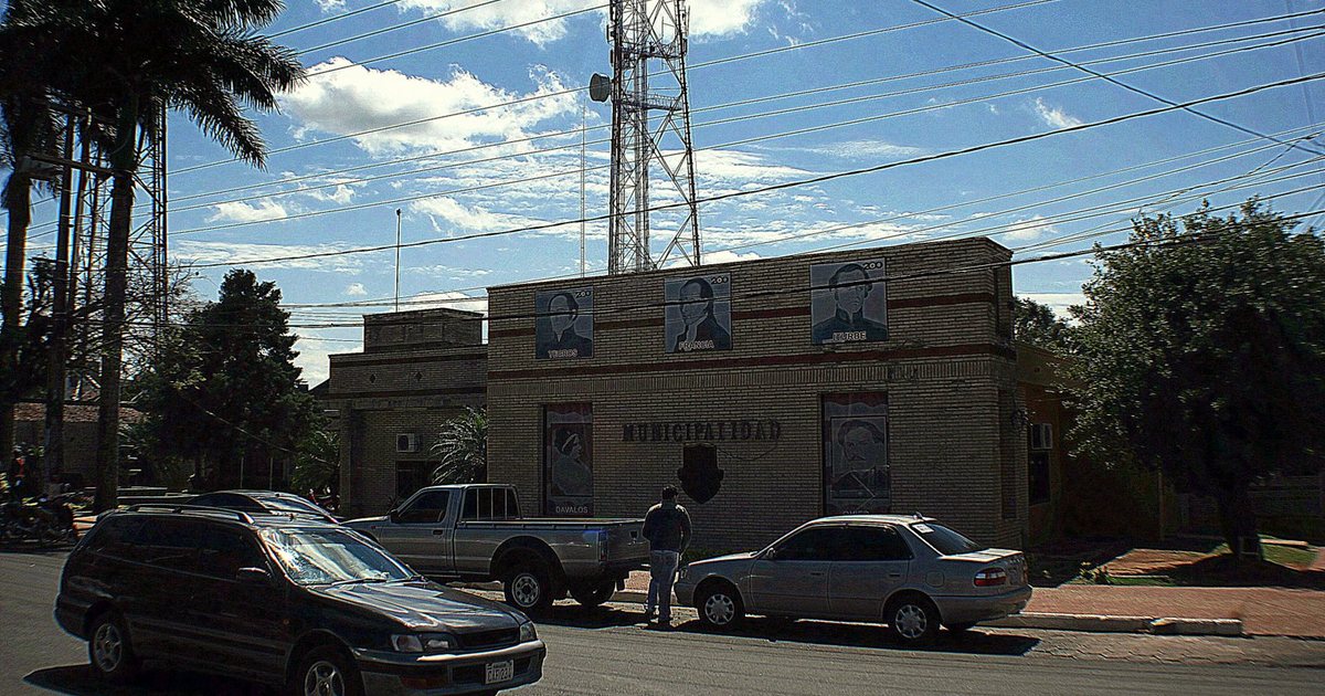 El edificio de ladrillo de la Municipalidad de Coronel Oviedo, con retratos de próceres en la fachada, una antena detrás y autos estacionados enfrente
