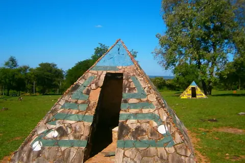 Pirámide de piedra con franjas verdes y una abertura, sobre un campo de pasto, con árboles y otra pirámide al fondo