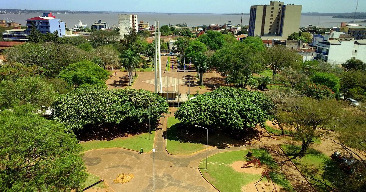 Vista aérea de la Plaza de Armas de Encarnación, con árboles frondosos, un monumento de columnas blancas, mástiles con banderas, edificios y el río Paraná al fondo