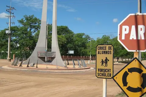 Rotonda de tierra con un monumento de hormigón en Filadelfia, junto a un cartel de pare y otro de cruce de alumnos en castellano y alemán