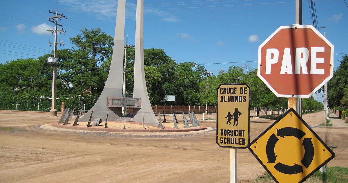 Rotonda de tierra con un monumento de hormigón en Filadelfia, junto a un cartel de pare y otro de cruce de alumnos en castellano y alemán