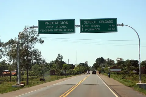 Ruta asfaltada con carteles verdes sobre la calzada que anuncian la zona urbana de General Delgado, entre árboles y campo