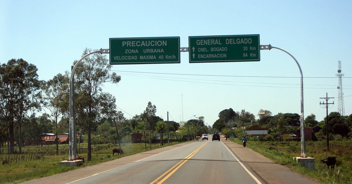 Ruta asfaltada con carteles verdes sobre la calzada que anuncian la zona urbana de General Delgado, entre árboles y campo