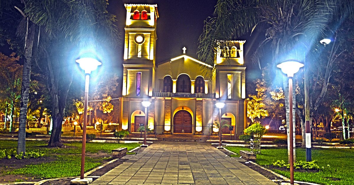 Iglesia de Guarambaré iluminada de noche al fondo de la plaza Emiliano R. Fernández, con faroles y palmeras