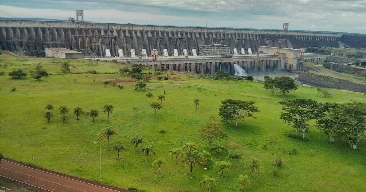La represa de Itaipú vista desde la margen paraguaya, con palmeras y césped al pie del muro y el vertedero al fondo