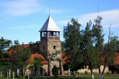 Iglesia de Colonia Independencia, con torre de piedra y campanario, techos de tejas rojas y árboles delante
