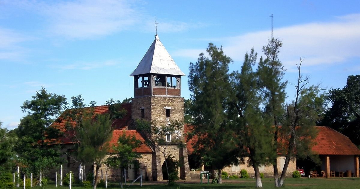 Iglesia de Colonia Independencia, con torre de piedra y campanario, techos de tejas rojas y árboles delante