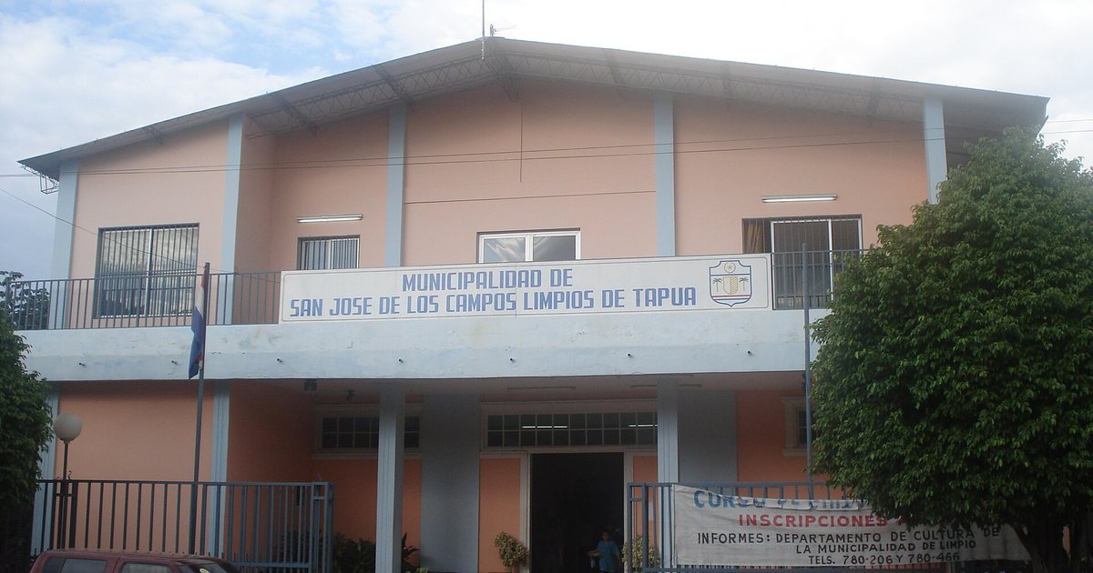 La sede de la Municipalidad de Limpio, un edificio de dos plantas con el cartel de la Municipalidad de San José de los Campos Limpios de Tapuá, una reja azul y un árbol a la derecha