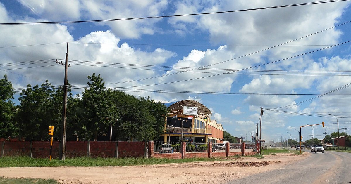 Sede de la Municipalidad de Loma Plata, con techo curvo y banderas, detrás de un muro de ladrillos junto a una avenida