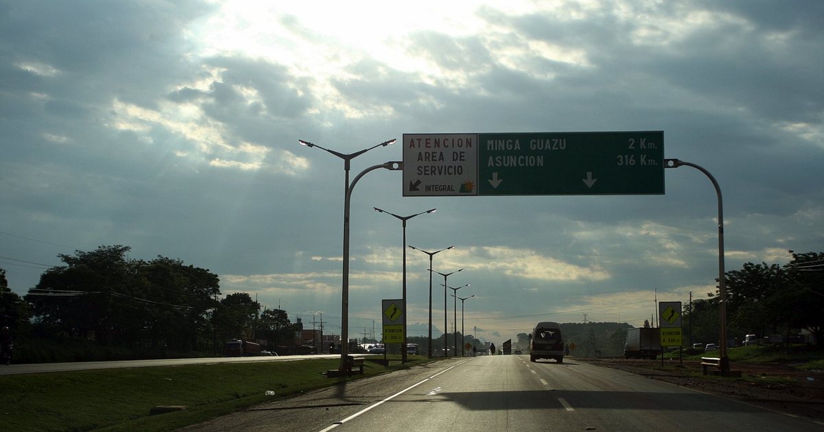 La ruta de entrada a Minga Guazú bajo un cielo nublado, con el cartel verde que marca 2 kilómetros hasta la ciudad y 316 hasta Asunción