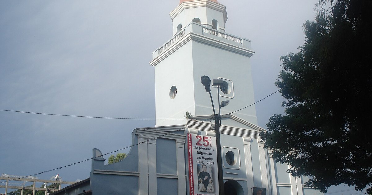 La torre blanca de la iglesia de Ñemby bajo un cielo nublado, con un farol en primer plano y un árbol a la derecha