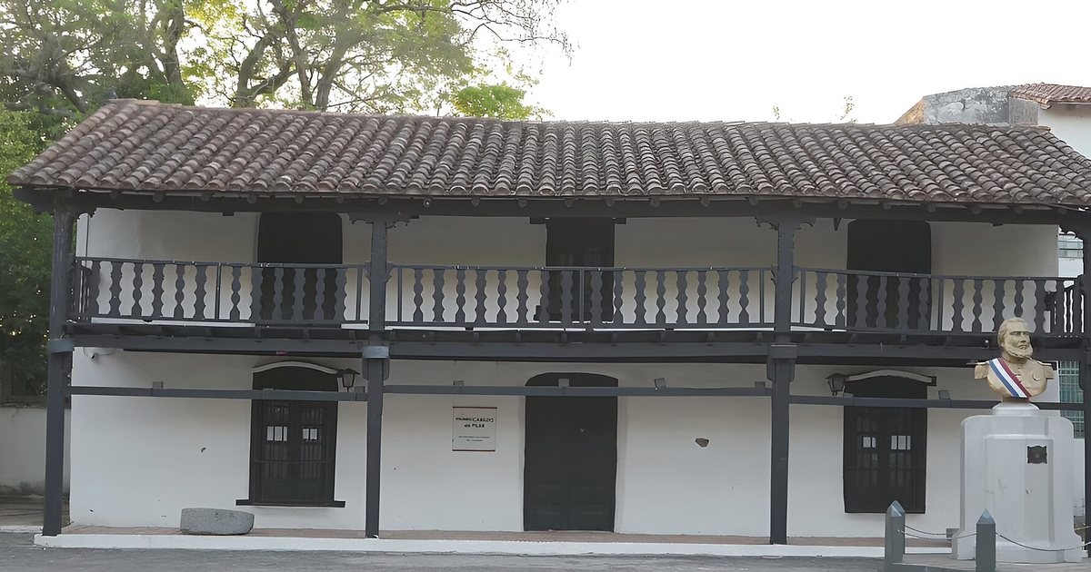 Casona colonial blanca del primer cabildo de Pilar, con techo de tejas, galería de madera oscura con balcón y un busto sobre un pedestal frente a la fachada