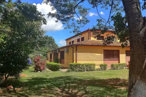 Edificio municipal de dos plantas pintado de amarillo, con techo de tejas, entre árboles y un jardín con arbustos