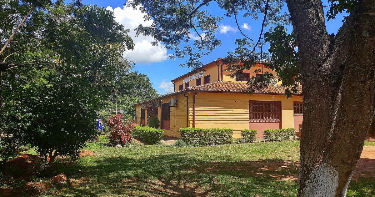 Edificio municipal de dos plantas pintado de amarillo, con techo de tejas, entre árboles y un jardín con arbustos
