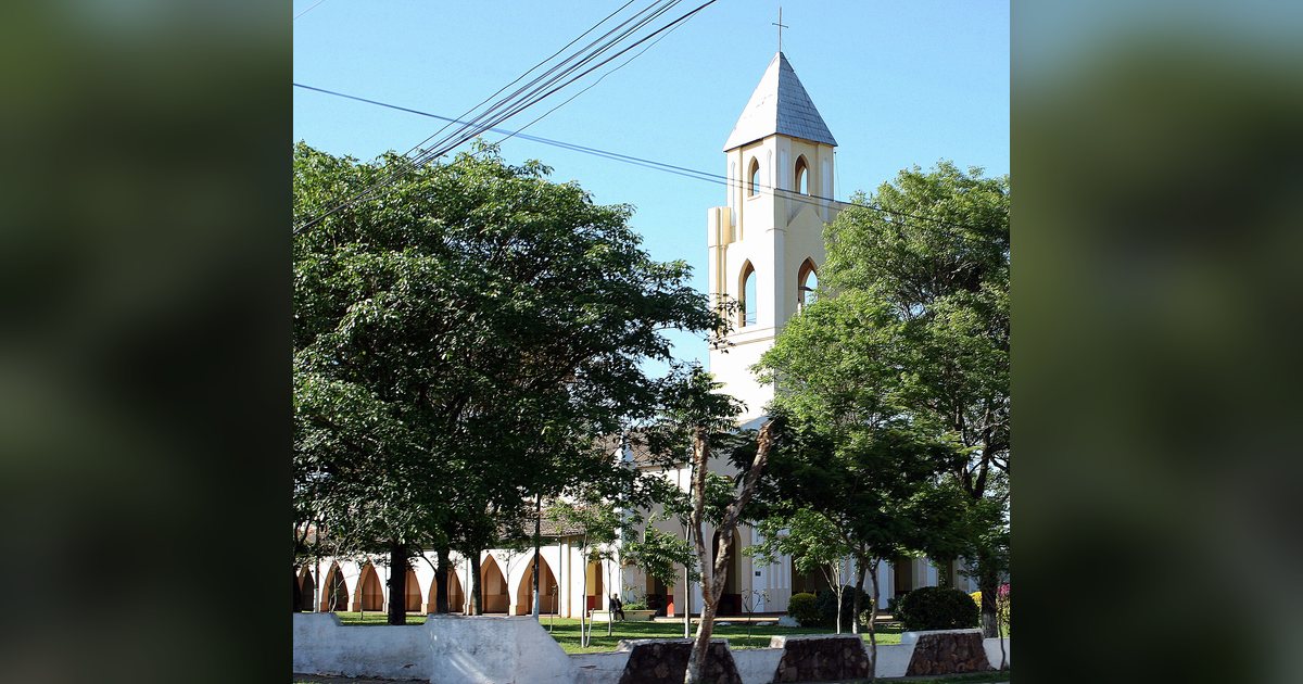 Iglesia blanca con campanario de techo a dos aguas y galería de arcos, detrás de árboles frondosos
