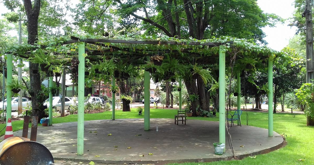 Pérgola de columnas verdes cubierta de enredaderas y helechos colgantes en una plaza arbolada de Salto del Guairá