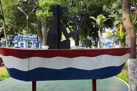 Monumento a los pescadores en la Plaza Central de San Antonio: una canoa pintada con los colores de la bandera paraguaya y la silueta negra de un pescador con su caña, entre árboles