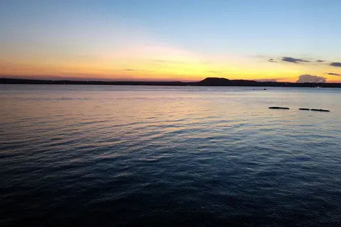 Atardecer sobre un lago en calma, con un cerro oscuro en el horizonte y el cielo naranja y azul