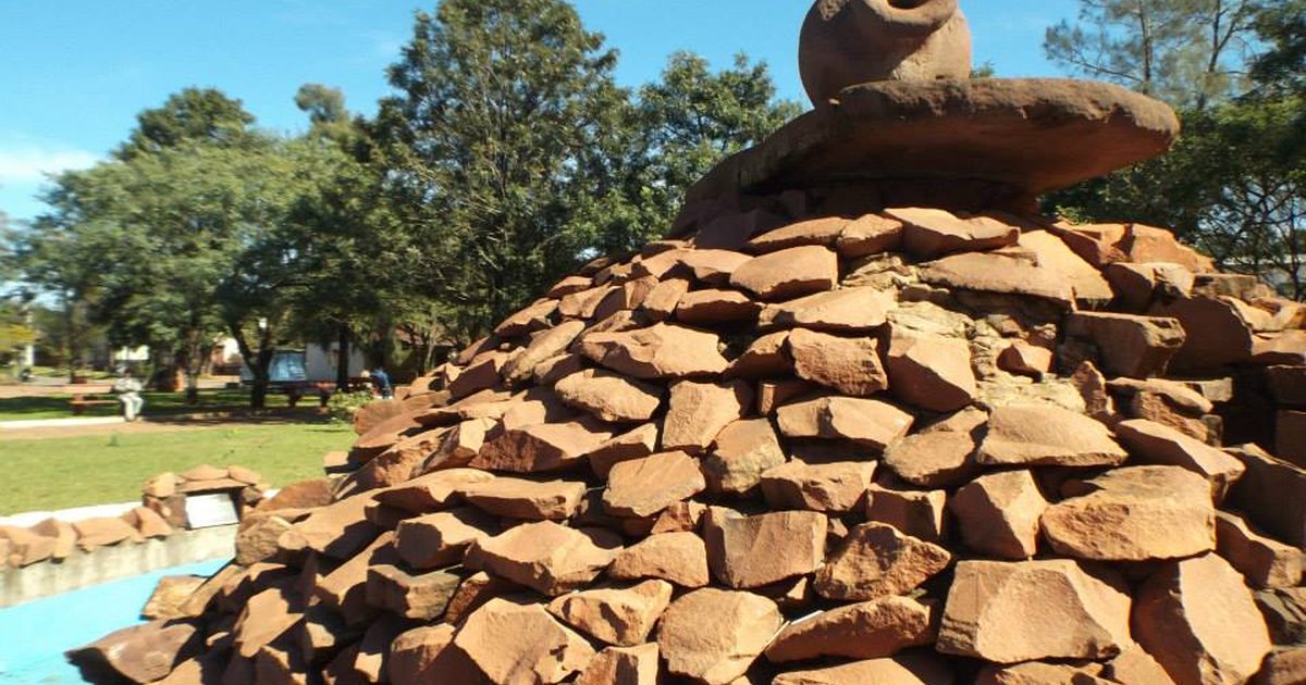 Escultura de piedras rojizas coronada por un cántaro de barro, en la plaza Boquerón de San Juan Bautista, con árboles y cielo azul