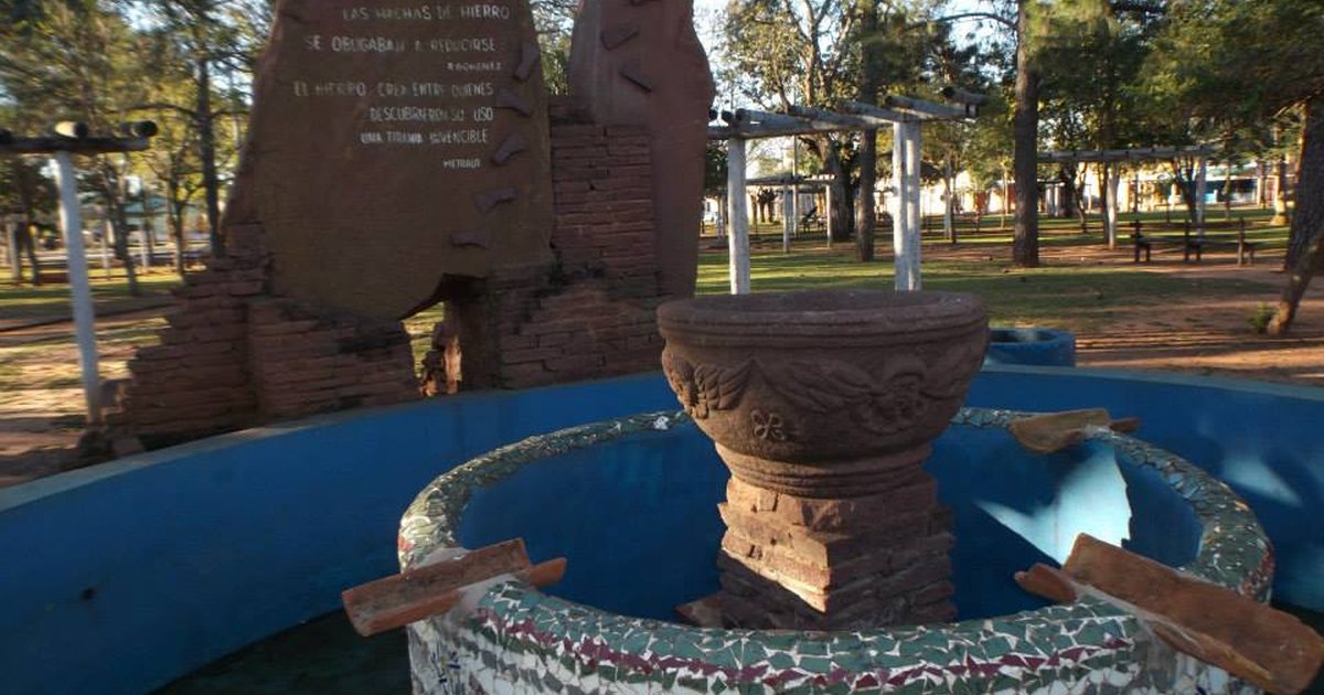 Fuente con mosaicos de colores y una pila de piedra tallada frente a un monumento de piedra con una inscripción, en una plaza arbolada