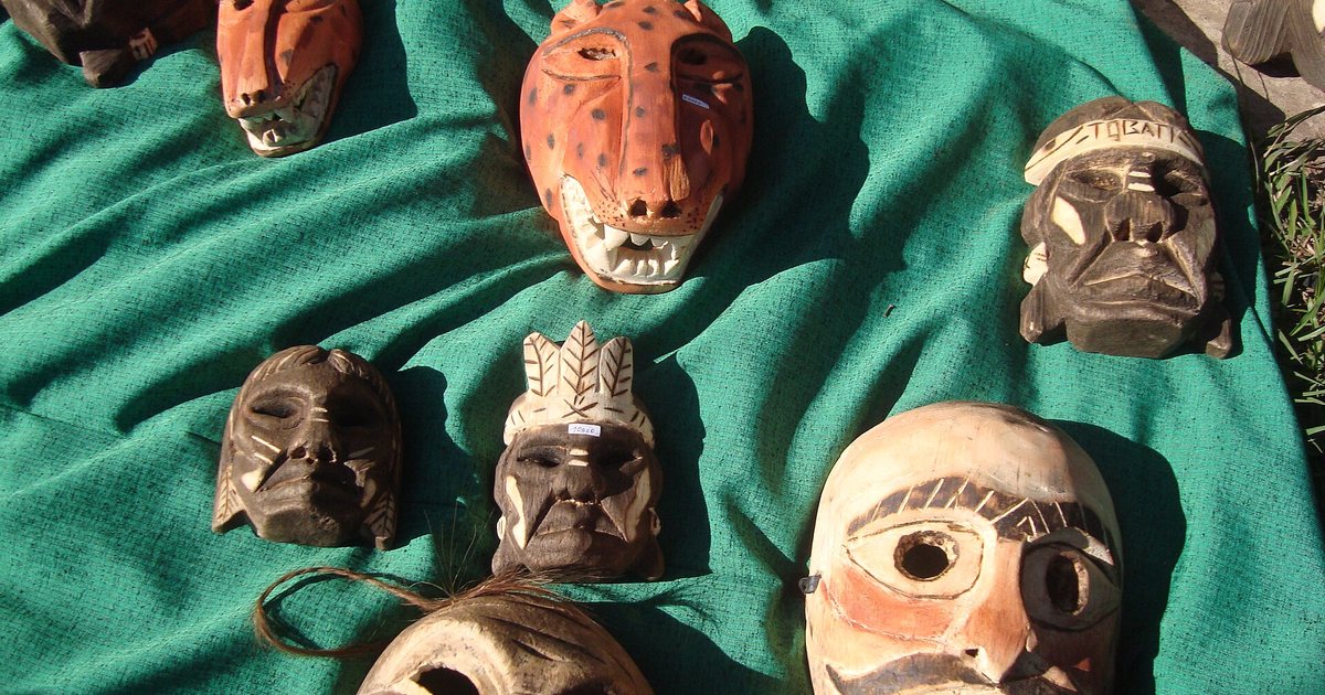 Máscaras de madera talladas y pintadas, entre ellas una de jaguar y una con bigote, sobre una tela verde en Tobatí