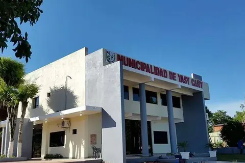 Edificio blanco de dos plantas con columnas y el letrero de la Municipalidad de Yasy Cañy, rodeado de árboles bajo un cielo azul