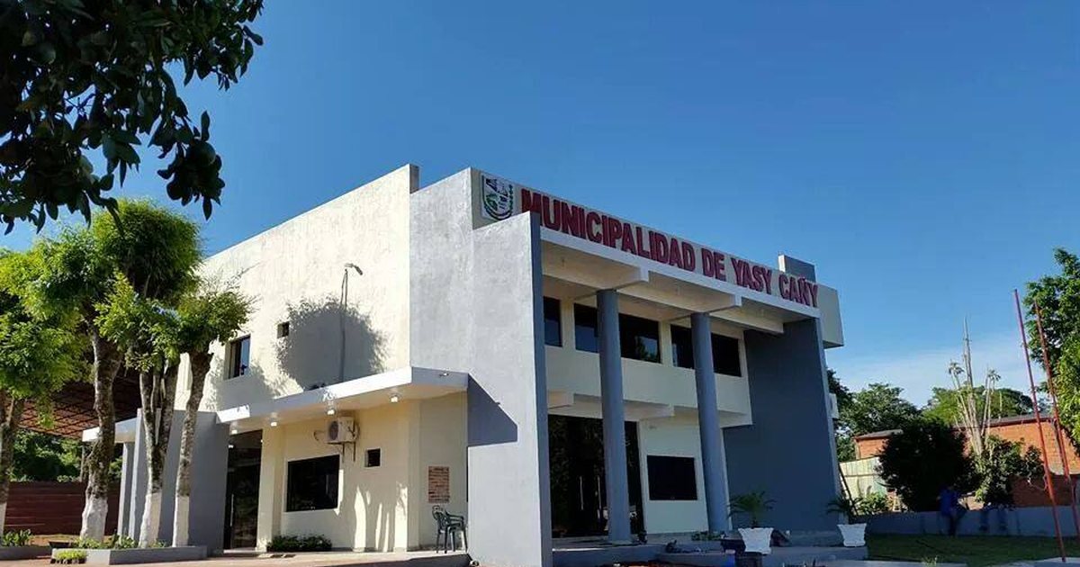 Edificio blanco de dos plantas con columnas y el letrero de la Municipalidad de Yasy Cañy, rodeado de árboles bajo un cielo azul