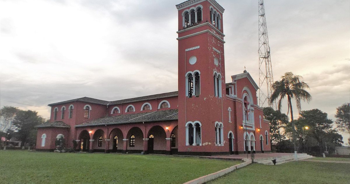 Iglesia de muros rojizos con arcos blancos y un campanario alto, rodeada de césped, bajo un cielo nublado al atardecer