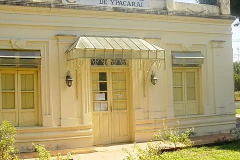 Fachada amarilla de la Municipalidad de Ypacaraí, con puertas de madera, un toldo de chapa y un jardín delante
