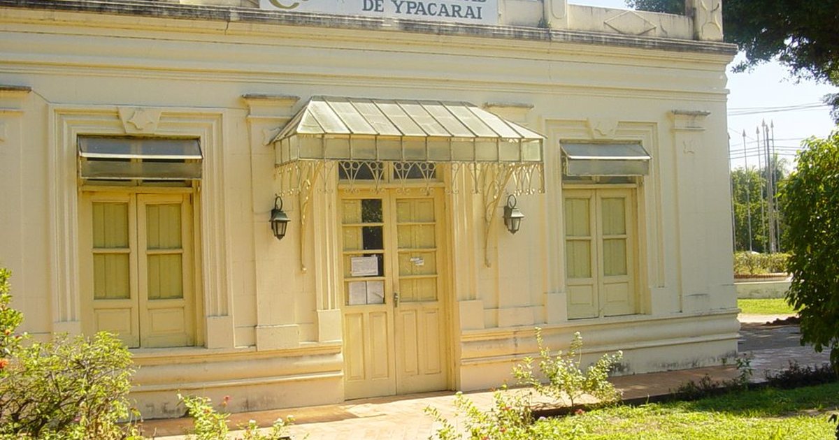 Fachada amarilla de la Municipalidad de Ypacaraí, con puertas de madera, un toldo de chapa y un jardín delante