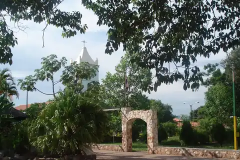 Plaza de Ypané con un arco de piedra, palmeras y árboles, y la torre blanca de la iglesia al fondo