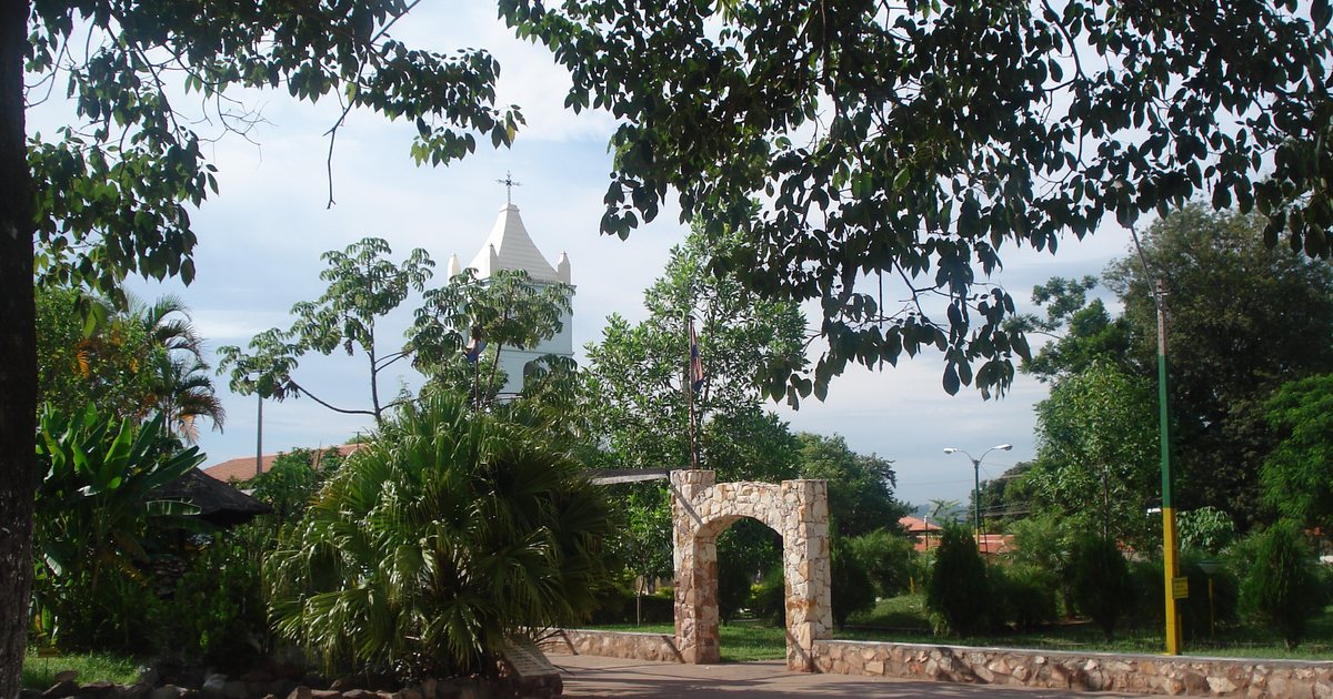 Plaza de Ypané con un arco de piedra, palmeras y árboles, y la torre blanca de la iglesia al fondo
