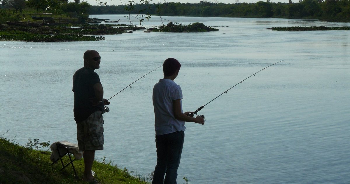 Dos personas pescan con caña desde la orilla del río Manduvirá, en Puerto Naranjahai, Paraguay